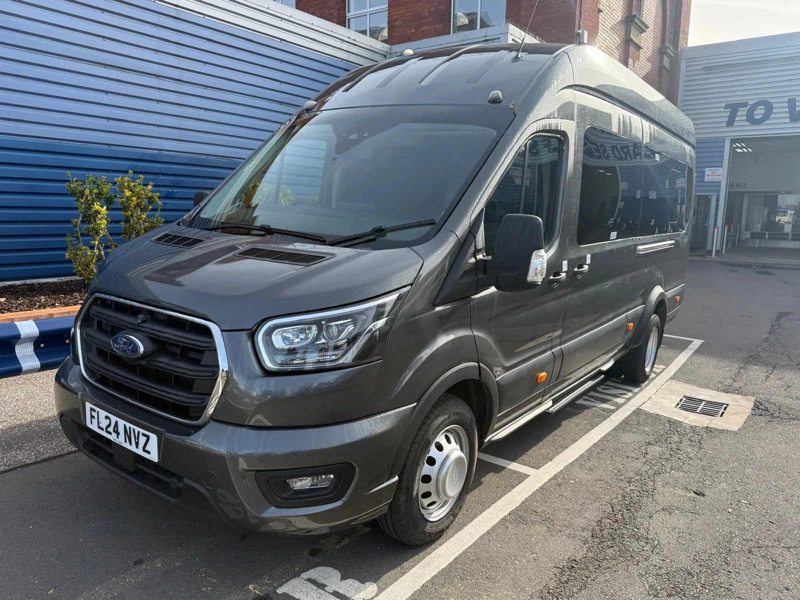 Grey 16 seater Ford Transit minibus parked in a marked bay, viewed from the front three-quarter angle