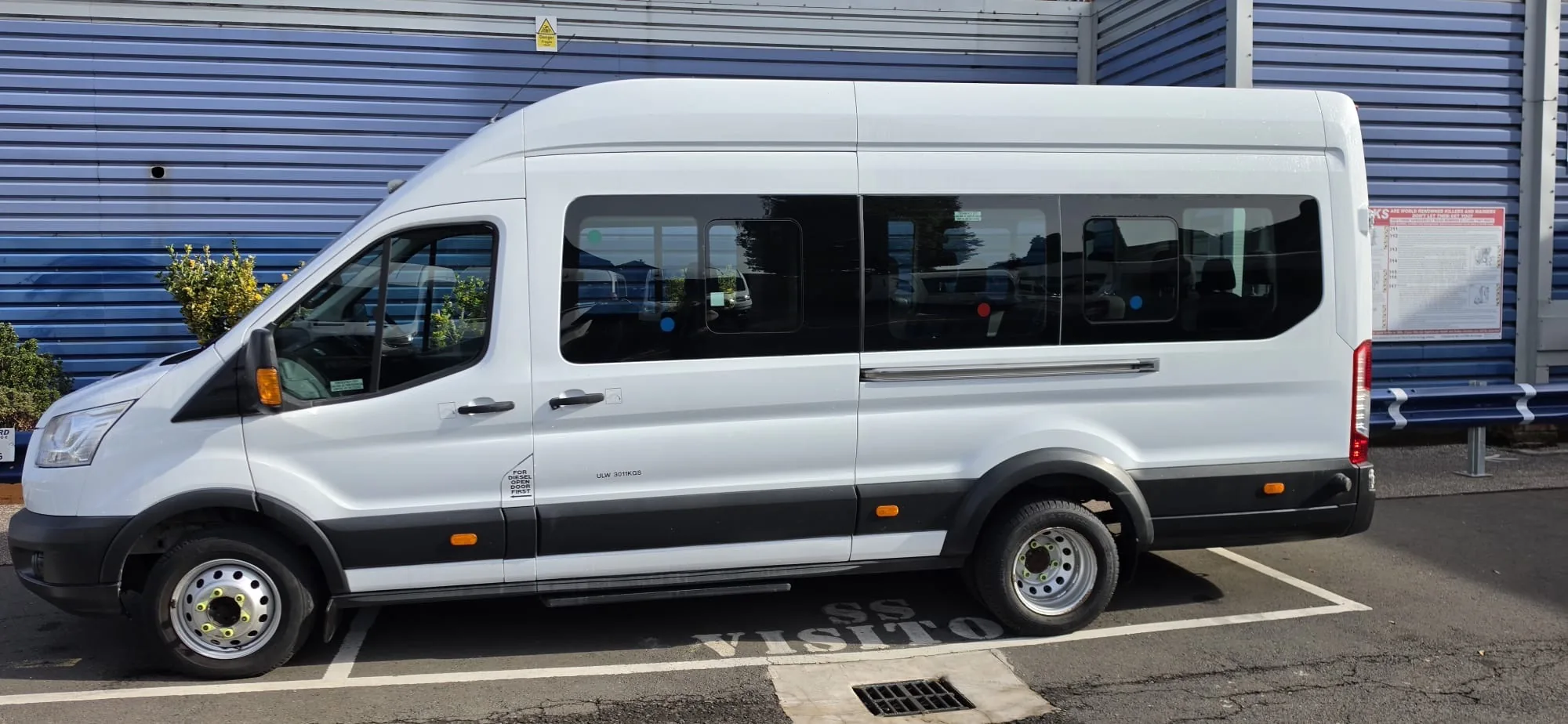Side profile of a long white 16 seater Ford Transit minibus with tinted windows, against a blue fence