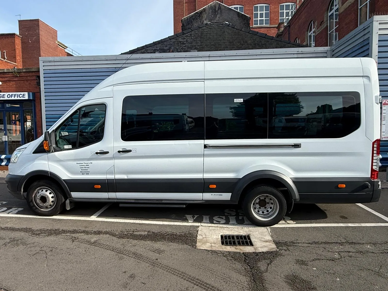 Side profile of a long white 17 seater Ford Transit minibus parked in front of a red brick building