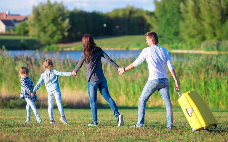 Family walking hand in hand towards a lake, the father pulling a yellow holiday suitcase