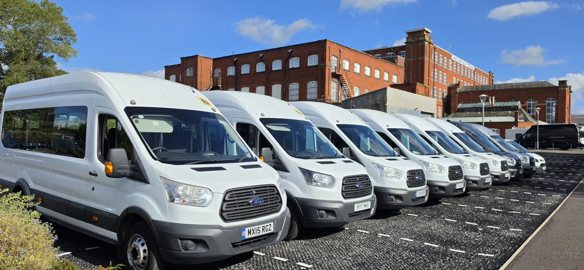 Row of white Ford Transit minibuses parked side by side on a sunny day in front of a red brick mill
